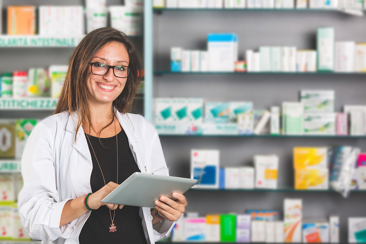 smiling female pharmacist on ipad in front of pharmacy shelves